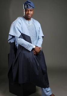 Femi Adebayo seated in a light blue agbada with a navy panel, a matching fila, and a silver wristwatch.