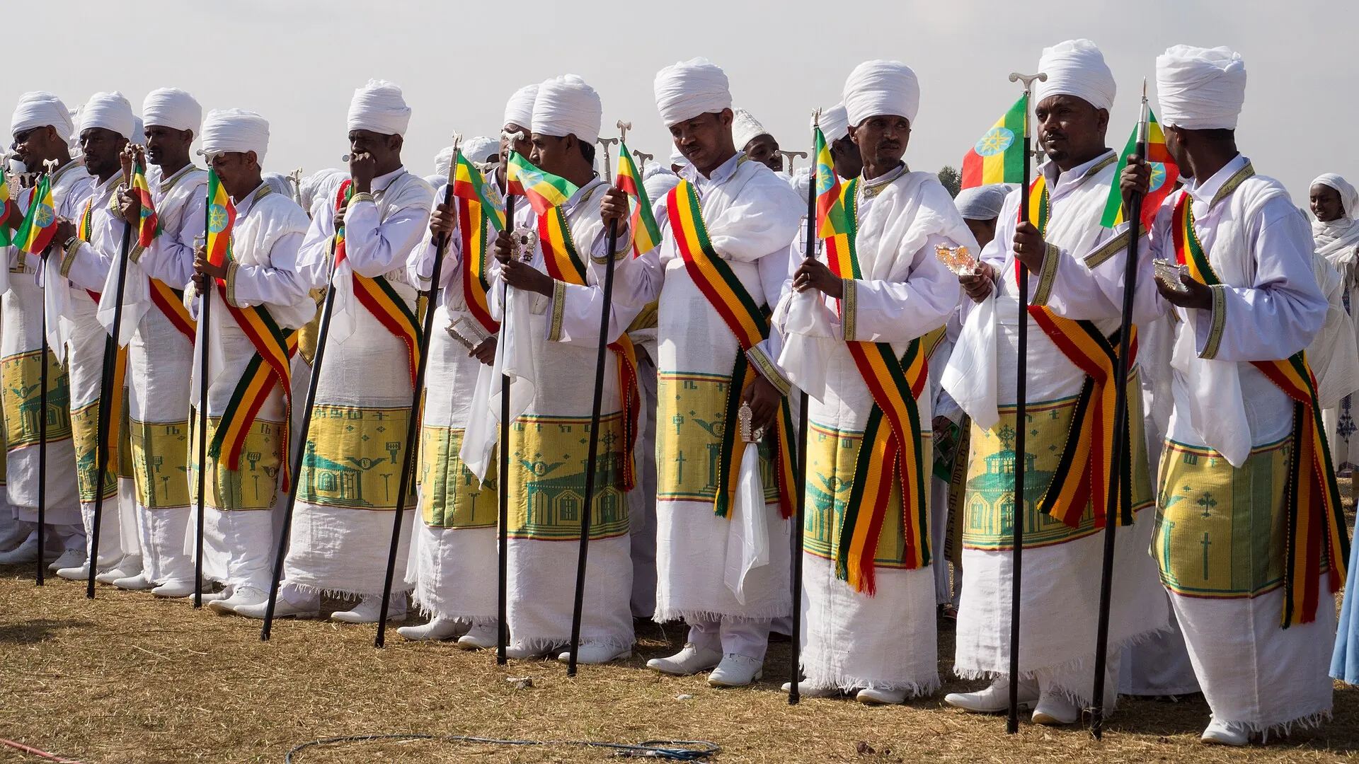 Young men at Timkat in Ethiopia in white robes and turbans, with green, yellow, and red sashes, holding flags and staffs.