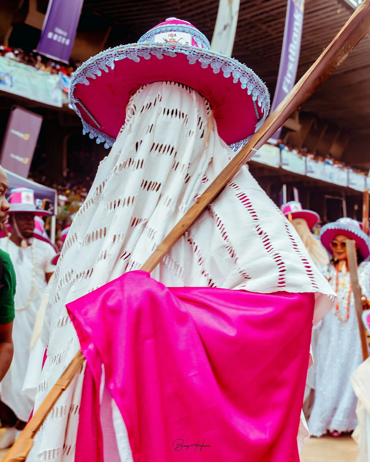An Eyo masquerader in Lagos in a white agbala and a wide red and white hat, holding a staff.