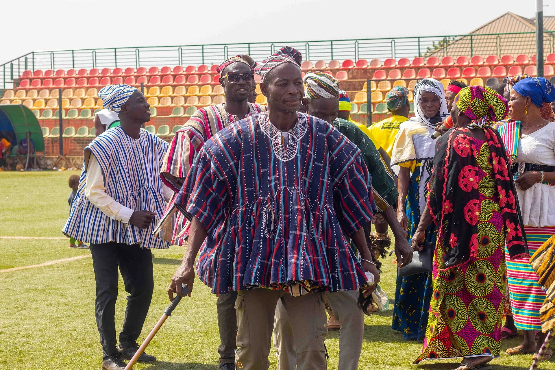 A Ghanaian chief in a striped batakari smock and cap walks with a staff at a durbar, attendants in cloth around him.