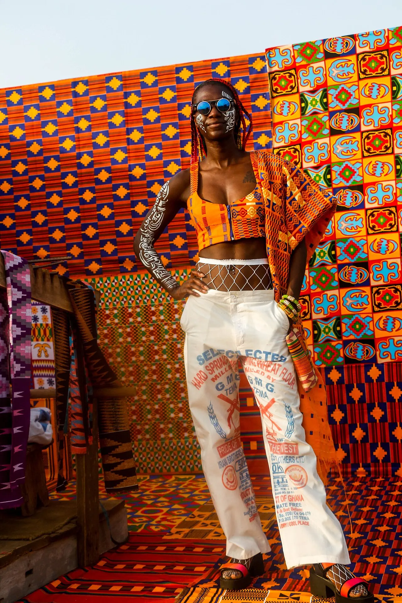 A woman in a kente top and white trousers, standing before a wall of woven kente in Ghana.