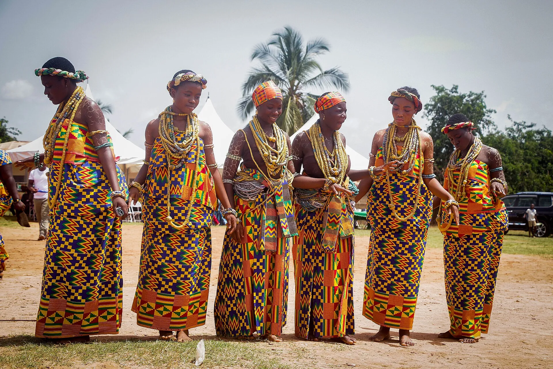 A line of women in Ghana in matching kente wraps and headbands, standing on red earth.
