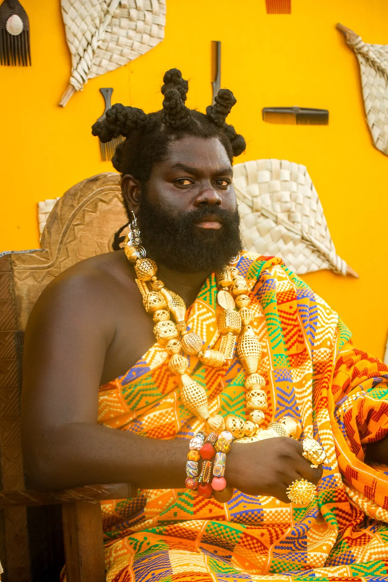 A man in Ghana dressed as a king, in kente cloth and gold jewellery, seated before a yellow wall.