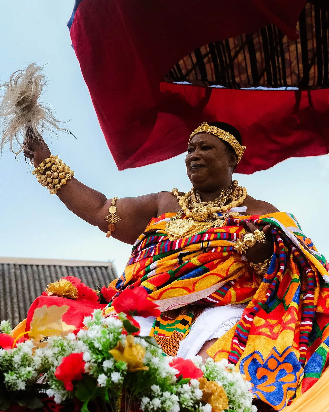 A Ghanaian queen mother in kente and gold jewellery, seated in state beneath a red umbrella.