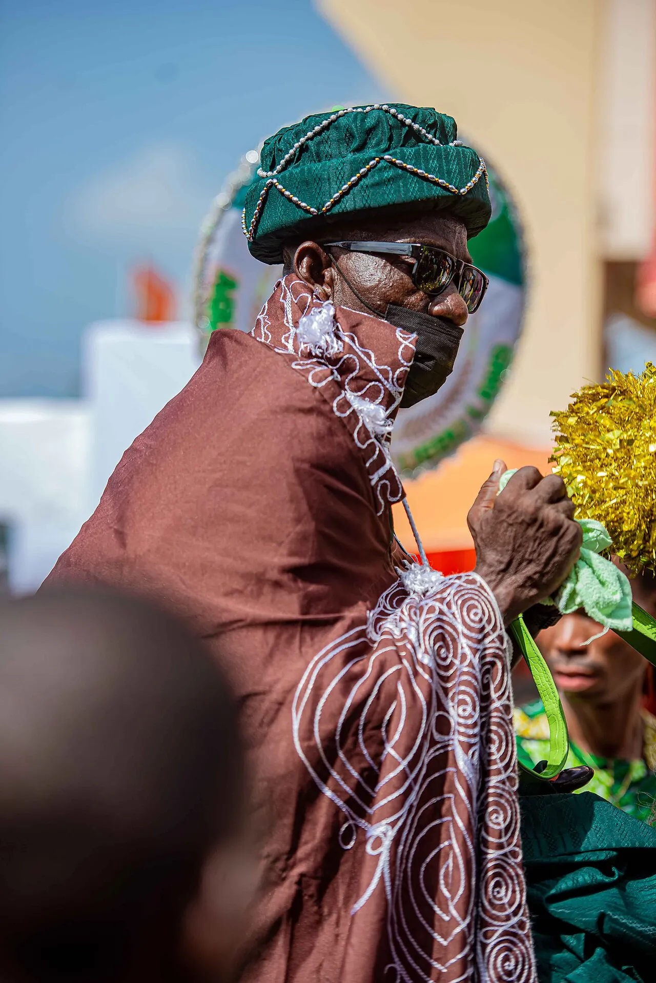 A man at Ojude Oba in Nigeria in a brown patterned agbada and a green beaded cap, his face veiled with beads.