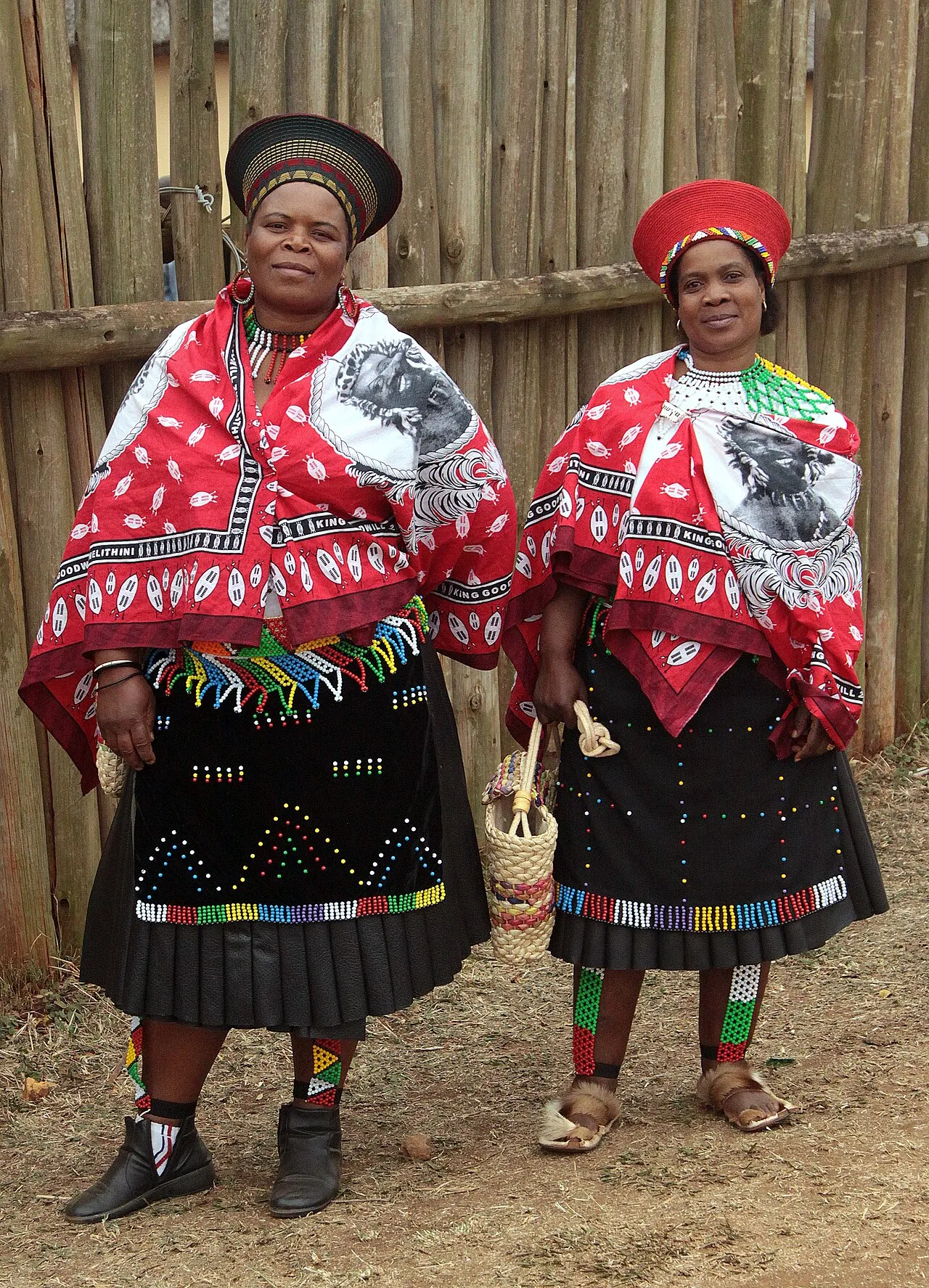 Two Zulu women in South Africa in red beaded capes, black pleated skirts, and wide ceremonial hats.